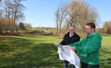 Dezernent Gerd Holzwarth mit Projektleiter Benjamin Kächele (r.). Im Hintergrund die gerodete Fläche am Bach, wo die Brücke hink