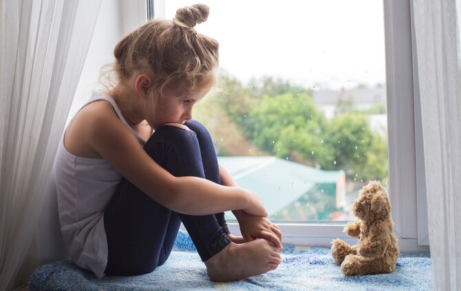 Sad litttle girl sits on a window sill and looks at a bear