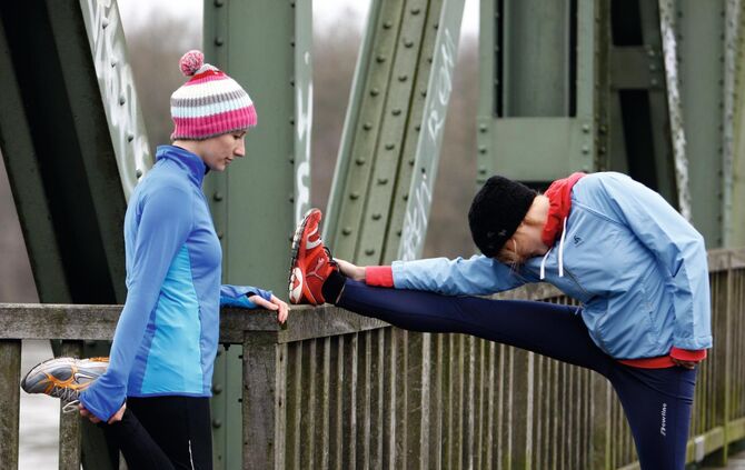 Zwei junge Frauen laufen im Winter, Dehnungsuebungen nach dem Lauf. Joggen in der kalten Jahreszeit, mit waermender, Wind- und Wasserdichter Funktionsbekleidung.