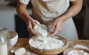 Baker pouring sourdough starter into flour in wooden bowl for baking bread