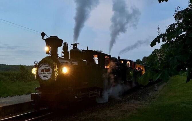 Eine historische Dampflokomotive fährt in der Dämmerung mit leuchtenden Scheinwerfern und Rauchfahnen über die Gleise.